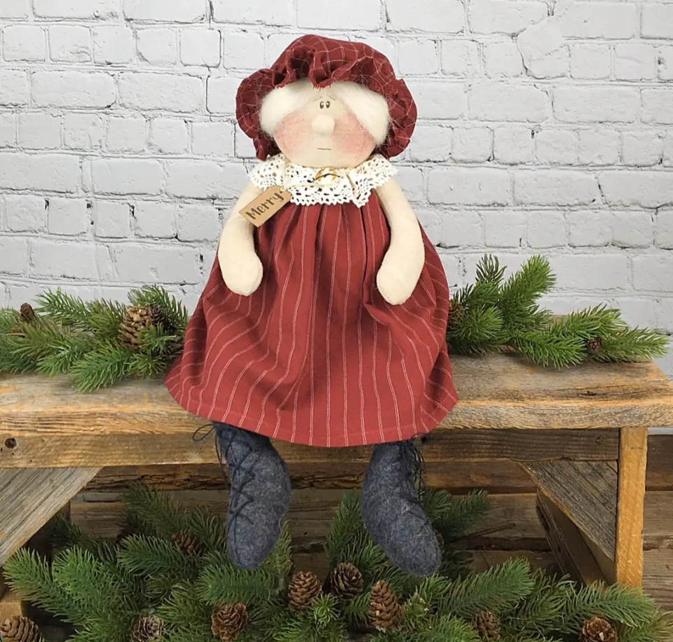 Whimsy Mrs. Claus doll named Merry sitting on a rustic wooden bench among pine branches, wearing a red striped dress, red plaid bonnet, lace collar, and gray boots