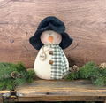 Primitive snowman named Winter with a floppy black hat, green plaid scarf, rusty bells, and a hand-lettered tag, sitting on a rustic wooden bench among pine branches