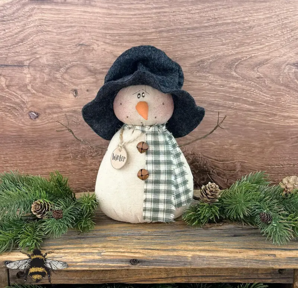 Primitive snowman named Winter with a floppy black hat, green plaid scarf, rusty bells, and a hand-lettered tag, sitting on a rustic wooden bench among pine branches
